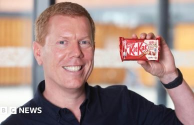 A smiling man with short ginger hair and blue eyes holds up a KitKat bar. He is wearing a navy shirt and black wrist watch. The background behind him is blurred.